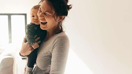 Close up of a smiling woman standing in her home carrying her child. Woman holding her baby and laughing standing in room.